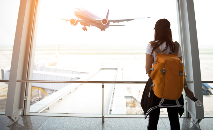 woman with orange backpack facing large window at airport - plane taking off - home