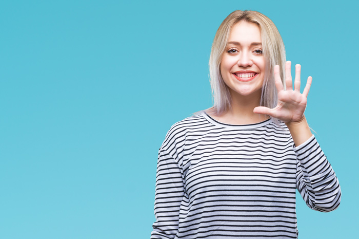 happy young woman holding up hand to show number five on sky blue background - intervention
