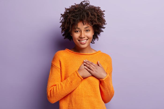 beautiful smiling African American woman in bright orange sweater with hands over heart on light purple background - gratitude