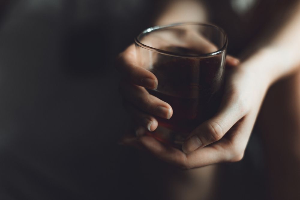 Close-up of a person holding a glass of dark alcohol in both hands, with a dim, moody background.