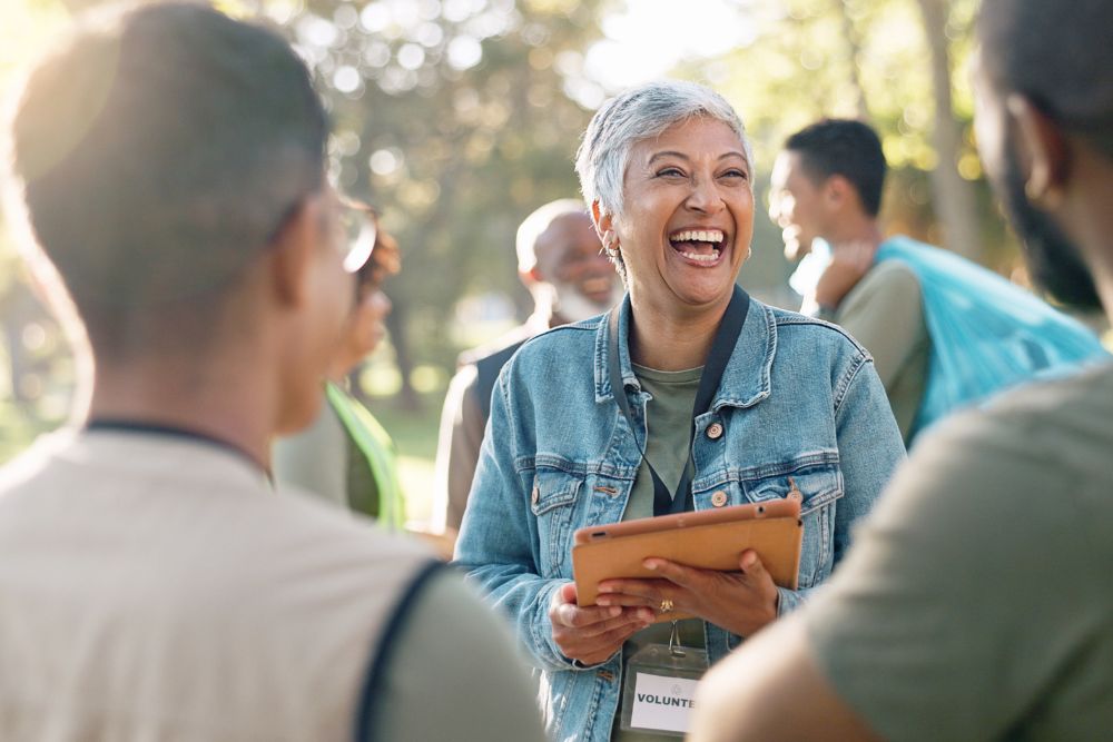 A cheerful woman wearing a volunteer badge and holding a tablet laughs while talking with a group of people outdoors. Others in the background are also smiling and engaged in conversation.