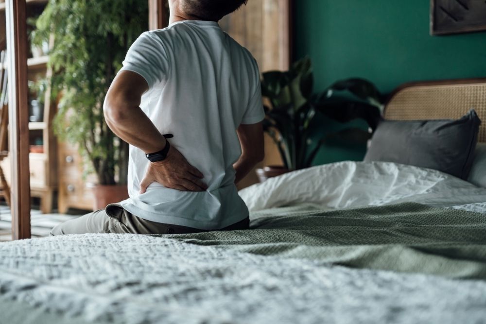 Person sitting on the edge of a bed holding their lower back in pain, symbolizing the struggles of chronic pain and its impact on daily life.