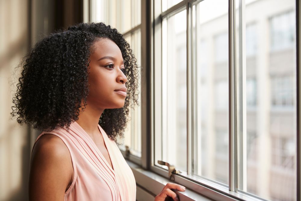 A woman stands by a window, looking outside with a thoughtful expression.