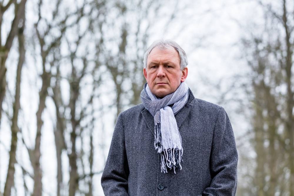 An older man stands outdoors on a cold, overcast day, wearing a gray coat and scarf. He looks serious and contemplative, with bare winter trees behind him.