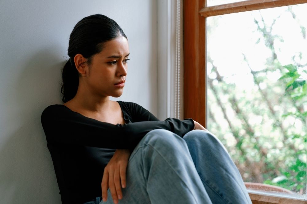 A young woman sitting on the floor by a window, knees pulled close, looking distressed and deep in thought.
