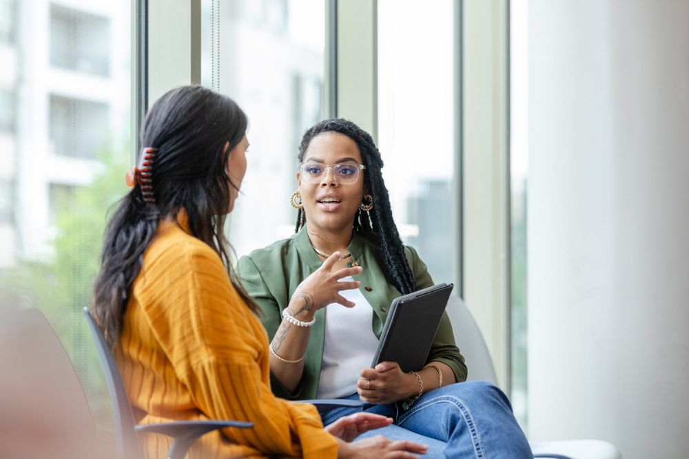 A therapist or counselor speaking with a woman in a bright office, holding a tablet while engaging in a supportive conversation.