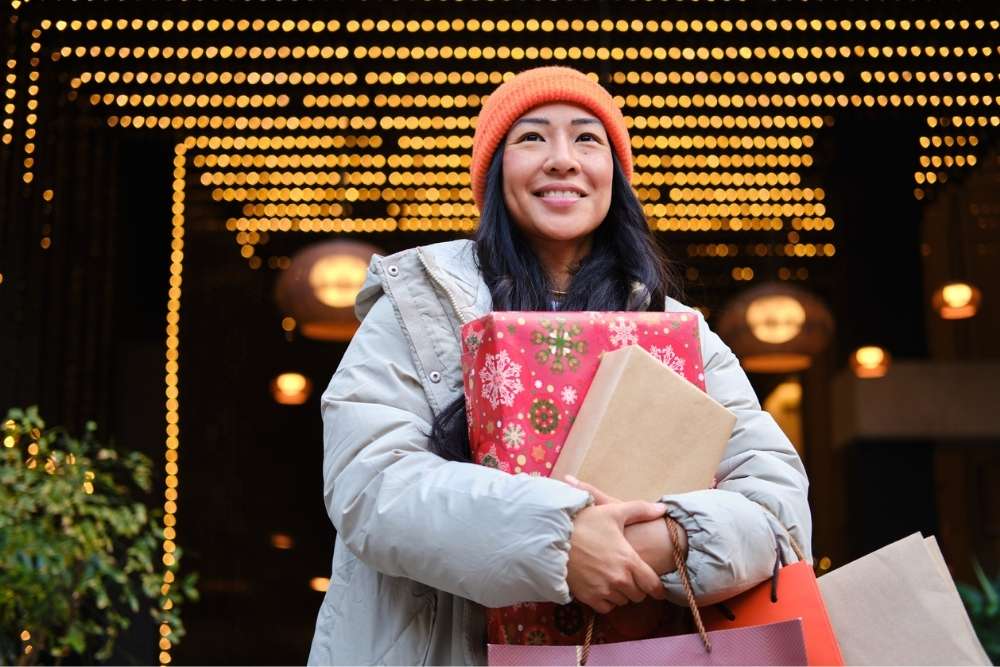 A smiling woman in winter clothing holds holiday shopping bags and wrapped gifts while standing under festive lights outdoors.