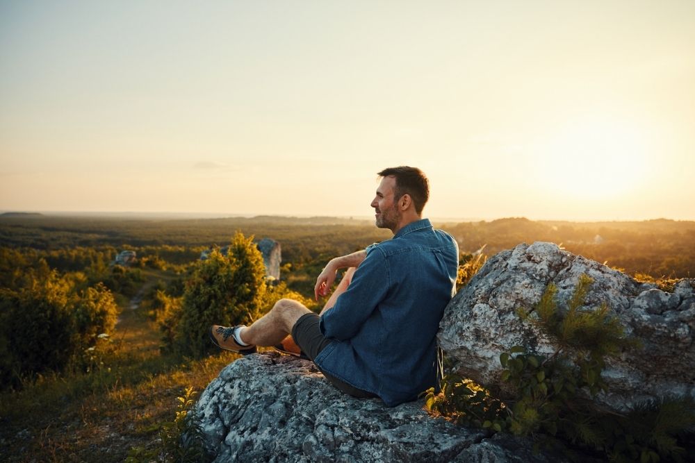 Person sitting on a rocky overlook at sunset, quietly reflecting while looking out over a wide natural landscape.