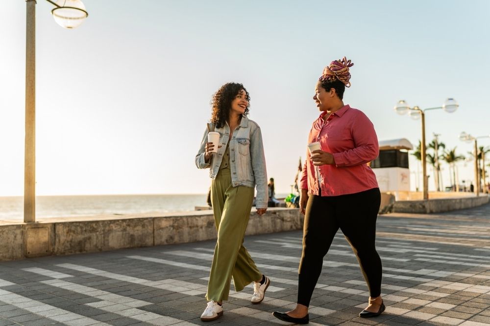 Two women strolling on a seaside promenade, laughing and holding coffee. The sun sets, casting a warm glow, and palm trees line the path. The photo depicts the characteristics of a good sponsor.